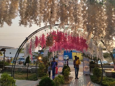 The image depicts a vibrant outdoor scene in Kondamaripalle, India. A decorative archway adorned with white and pink flowers frames a walkway paved with patterned tiles. A family, consisting of a woman in a blue sari, a young boy, and a man carrying a child in a yellow dress, is walking through the archway. Adjacent to the walkway are green bushes and trees. Behind the archway, there are buildings, parked cars, and other individuals in the background. The sky is a light color, suggesting either early morning or late afternoon.