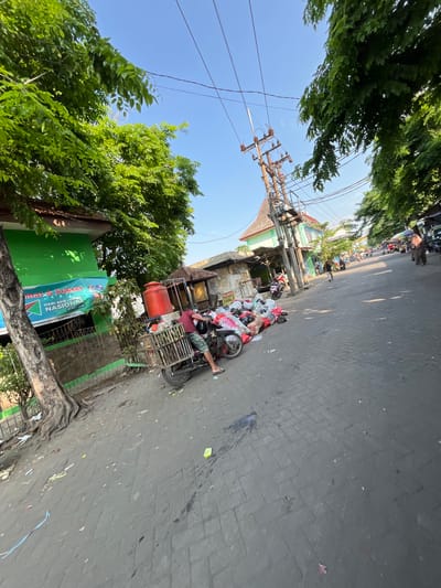 The image depicts a street scene in Juwet Kenongo, Indonesia, with a clear blue sky and lush green trees lining the road. In the foreground, a man is operating a two-wheeled cart loaded with refuse, which is parked next to a collection of garbage bags and assorted items. The cart is equipped with a large red water tank. To the left of the cart, a green building with a banner displaying text in Indonesian can be seen. The street itself is paved with cobblestones, and several motorcycles and a few other people are visible further down the road, suggesting a bustling urban environment. Power lines and a utility pole are prominent in the upper portion of the image. The scene appears to be captured during daylight hours, possibly in the morning or late afternoon, given the quality of light and the shadows.