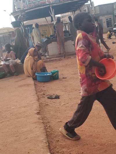 This is a daytime, full-shot image depicting a street scene in Funtua, Nigeria. The foreground is dominated by a young Black boy walking from right to left, wearing a vibrant, patterned long-sleeved shirt and dark trousers. He holds an orange bowl in his right hand. To the left of the boy, a woman wearing a yellow hijab and gown sits on a curb, with her back to the camera. Next to her are two buckets, one blue and one green, on the ground. Further back, other individuals are visible, some standing and some sitting, near market stalls. The environment appears to be a dusty, unpaved street with a building structure providing shade behind the market stalls. The overall impression is one of a busy, everyday urban environment.