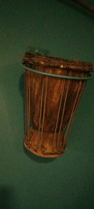 A single, dark-toned, goblet-shaped wooden object is prominently displayed against a plain, dark forest green background. The item, originating from Yola, Nigeria, features a rustic and aged appearance. Its body is constructed from numerous distinct vertical wooden staves or slats, estimated at 15-20 visible, which are held together by horizontal bands. A slender, turquoise-colored band encircles the upper portion of the object's body. The top edge presents a thick, dark, rounded rim, exhibiting several small, light-yellow circular markings, potentially decorative tacks or structural elements. The base of the object is wider and flared. The wood displays varied dark brown to reddish-brown tones with visible grain and natural imperfections. Soft, indirect frontal lighting casts subtle shadows to the object's right and lower side on the uniform background. No individuals, other subjects, actions, or interactions are present in the scene.