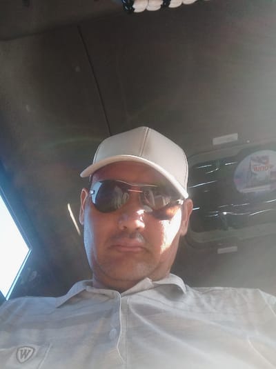A medium shot photograph shows a man wearing a light-colored baseball cap and sunglasses, looking directly at the camera. He has a mustache and is wearing a light-colored collared shirt with a logo on the left breast. The background appears to be the interior of a vehicle, with part of a window on the left and a reflective surface with text and images on the right. The lighting suggests it is daytime, with sunlight creating highlights on the man's face and shirt. The location is identified as Nukus, Uzbekistan.
