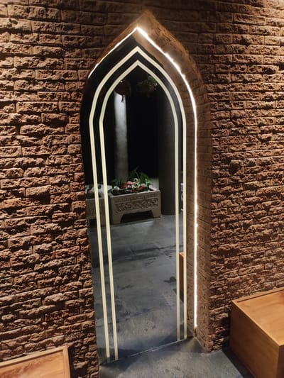 The scene, located in Mas, Indonesia, features a pointed archway set within a reddish-brown brick wall. The wall consists of rough, irregular bricks with visible mortar. The archway is illuminated by two parallel, glowing light strips along its inner contour. Through the archway, a dark grey tiled floor extends into a dimly lit interior space. In the mid-ground, a light-colored, rectangular stone planter with carved details contains green and reddish-pink foliage. Behind the planter, a dark cylindrical column rises, partially lit by an overhead light source. In the foreground, at the lower-left and lower-right, sections of light-colored wooden rectangular structures, possibly benches or boxes, are visible. No human subjects are present.