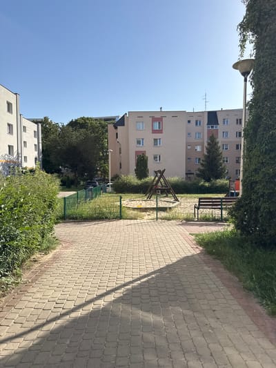 The image shows a courtyard between apartment buildings in Białystok, Poland. A paved walkway runs along the right side of the frame, bordered by green grass and bushes. In the center of the image, a small playground with a sandbox and a wooden swing set is visible, separated by a green metal fence. Behind the playground, a wooden bench is placed near a tall evergreen tree.
Several multi-story apartment buildings with light-colored facades and numerous windows dominate the background. One building has prominent red accents around some of its windows. The sky is clear and blue, indicating a sunny day. A lamppost is visible on the left side of the image, partially obscured by dense green foliage. The scene suggests a residential area designed with communal outdoor spaces for residents. The time of day appears to be late morning or early afternoon, given the angle of the shadows cast by the walkway and the trees. No people are visible in the image. The overall mood is calm and ordinary, depicting a typical urban residential environment on a pleasant day.