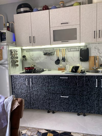 This is a medium shot, eye-level, indoor photograph of a kitchen with a microwave and refrigerator on the left, a stovetop and sink on the right, and cabinets above and below. The kitchen appears to be in a residential setting. The overall lighting is bright, with overhead lights illuminating the space. The lower cabinets are black with a floral pattern. The upper cabinets are white with a similar floral pattern. The kitchen is clean and organized, with various utensils and cookware visible. The photograph is taken from a slightly angled perspective, showing a portion of the floor and a small rug in the foreground. The location is Maksimkovo, Russia. There are no people in the image.