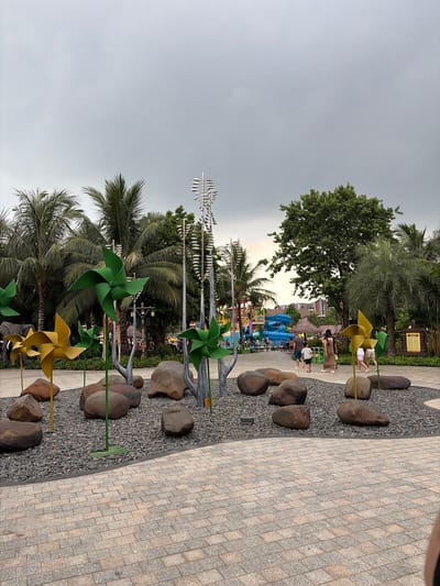 The image depicts an outdoor scene with a paved walkway in the foreground, made of light-colored rectangular tiles. Beyond the walkway is a landscaped area with dark gray gravel, large rounded rocks, and several decorative pinwheels in green and yellow. Palm trees and other lush foliage surround the area, with a hint of a water park with blue slides visible in the background. Several people are walking in the distance. The sky is overcast. The image was captured in Nghĩa Trụ Commune, Vietnam.