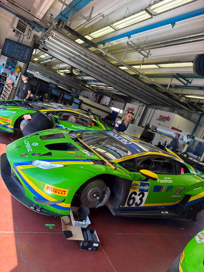 The image depicts the pit lane of the Mugello Circuit in Scarperia, Italy, a professional motorsport venue. Two vibrant green racing cars, likely Lamborghinis due to their distinct design and branding, are the primary subjects. The car in the foreground is positioned at an angle, with its front wheel removed and brake pads visible in a box on the ground, indicating maintenance or repair work is in progress. The car behind it is also being attended to by a mechanic.

Several individuals are present in the scene, some appearing to be drivers or pit crew. One person, wearing glasses and a black t-shirt, stands to the left, looking towards the monitors. Another individual, seated with a laptop, is positioned on the back of the second car. A man in a blue and white polo shirt is seen leaning over the front of the second car, possibly inspecting the engine. The environment is a typical racing garage, with overhead fluorescent lighting, exposed pipes and conduits, and various pieces of equipment. Monitors displaying race data are visible in the background, further confirming the racing context. The floor is a dark red epoxy, common in professional pit garages. The cars are adorned with numerous sponsor logos, including Pirelli, Lorenz, and Fastron, indicative of their professional racing status.
