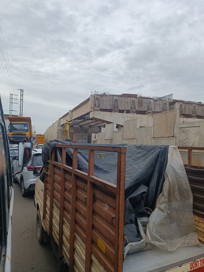The image captures a traffic jam on a cloudy day, likely in Balanagar, India, with ongoing construction of an elevated structure. In the foreground, a brown wooden truck bed is visible, partially covered with a black tarp and a white plastic sheet. Behind it, a light-colored truck is stopped. The road is lined with several cars, including a white SUV. Further ahead, a yellow dump truck is parked in the traffic. A golden statue of a person is positioned on the under-construction elevated road, pointing towards the right. The construction site features concrete walls and a superstructure with exposed rebar. In the background, electrical towers and overhead wires are present. The overall scene suggests a busy urban environment experiencing traffic congestion and significant infrastructure development.
