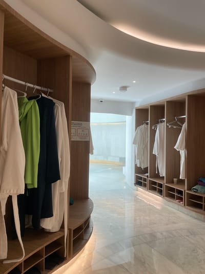 The image depicts a wooden shelving unit within a building, likely a spa or wellness center, containing folded towels, green and blue garments, and white robes on hangers. The shelving extends overhead, containing white robes on hangers. The surrounding architecture is characterized by a curved ceiling and marble-tiled floors. The language on a sign present in the shelving unit is Catalan. This suggests the location is within the Principality of Andorra, and most likely in the city of Les Escaldes, given the local use of Catalan language. There are no people visible in the image.