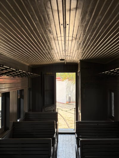 This image shows the interior of a passenger rail car, located in Cuautla, Mexico. The walls are made of horizontal wooden planks. A luggage rack runs along the top of the left wall. "DAMAS" is written on the wall above the doorway. The door leads to an outside scene of a building, trees, and ground. Text is printed on the wall near the door, which warns against the use of flammable materials. The number "993" is displayed above a window. Passenger seats are visible on the right.