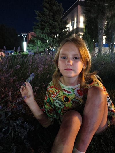 The image features a young girl sitting in a field of lavender at night. The girl has fair skin, blonde hair, and is looking directly at the camera with a neutral expression. She is wearing a colorful t-shirt with comic book-style graphics and the words "BOOM!", "WHIZ!", and "BANG!". Her right hand is holding a sprig of lavender. In the background, a modern building with illuminated windows is visible, surrounded by trees. A tall lamp post with a V-shaped top and a bright light is also present. The scene appears to be set in an outdoor urban or suburban area, given the presence of the building and lamp post, and the lavender suggests a park or garden setting. The overall lighting suggests it is nighttime, with artificial lights illuminating the scene. The presence of lavender indicates a temperate climate. The context provided states the location is Krinitsa, Russia.