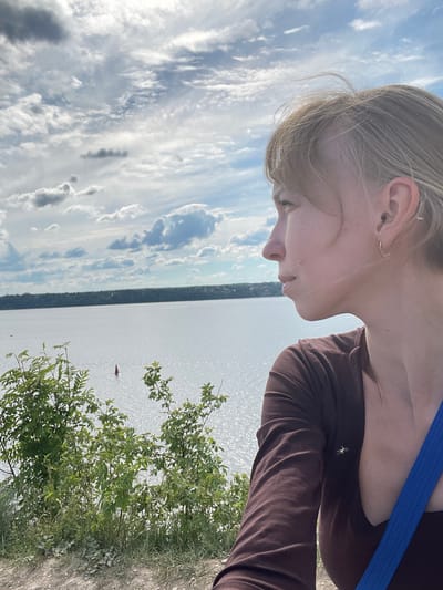 The image captures a profile view of a person looking out over a vast body of water from an elevated position. The subject, a person with light-colored hair, is seen from the chest up on the right side of the frame, facing left. They are wearing a long-sleeved brown top, with a bright blue strap visible on their right shoulder. Their gaze is directed downwards and outwards towards the scene.

The mid-ground features a wide expanse of water, likely a river or large lake, shimmering brightly under what appears to be sunlight. A small, red, indistinct object, possibly a buoy or a distant boat, is visible on the water's surface. In the far distance, a thin strip of land or shoreline is visible across the water. The background, particularly on the left and upper parts of the frame, is dominated by a sky filled with a mix of white and grey clouds, indicating a partly cloudy but bright daytime. In the lower right foreground, green foliage, consisting of bushes and smaller plants, frames the edge of what appears to be a bank or cliff.

The scene suggests an outdoor setting at "Пятый километр, Russia," likely a scenic overlook, where the person is engaged in observing the landscape. The weather is sunny with clouds, and the time of day is daytime. No discernible text is visible in the image.