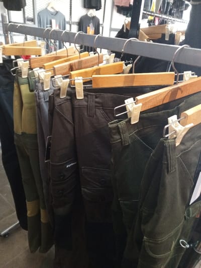 The image depicts an interior view of a clothing retail establishment in Baza, Myanmar (Burma), focusing on a display of multiple pairs of pants. A dark-colored horizontal metal clothing rack supports numerous wooden hangers, each featuring a metal hook and white plastic clips suspending a pair of trousers. The pants are primarily in utilitarian colors, including dark green, olive green, grey, black, and brown. Several pairs exhibit characteristics of cargo pants, with visible pockets on the thighs. One pair of dark brown cargo pants prominently displays the brand name "Explore" embroidered on a leg pocket. In the blurred background, additional clothing items, such as t-shirts in various colors, hang on other racks or are displayed on shelves, against a background suggesting metal grid fixtures. The floor below the display appears to be a light-colored tiled surface. No individuals are visible within the frame.