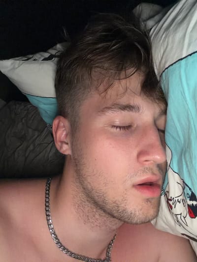 A young man with light brown hair and closed eyes is lying on his side, resting his head on a pillow. He is wearing a silver chain necklace. His mouth is slightly open, and he has some stubble on his face. The pillow has a blue and white striped pattern with a cartoonish drawing. The background is dark and appears to be a bedroom. There is no indication of the city of Vanino, Russia, in the image.