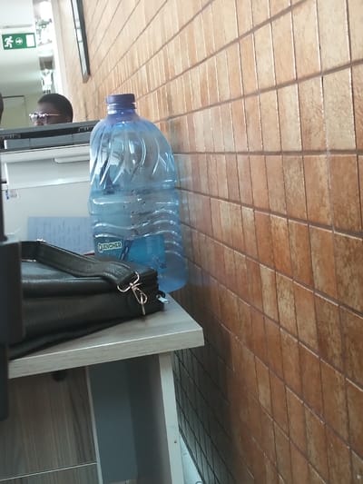 The image displays an indoor scene within Viwandani ward, Kenya, featuring a beige tiled wall on the right and an office-like setting on the left. In the foreground, a large, clear plastic "QUENCHER" water bottle with a blue cap and condensation is positioned on a light-colored desk surface. To its left, a black synthetic leather shoulder bag with a silver clasp rests on the same surface, partially obscuring a stack of papers with blue markings. Behind these items, a white office machine, possibly a printer or copier, is visible. Above and behind the office machine, the upper part of an individual's head, appearing to be of dark complexion and wearing glasses, is present. In the upper-left background, a green exit sign with a white running man pictogram and a right-pointing arrow is visible, reflected in a surface that also shows a black framed object.