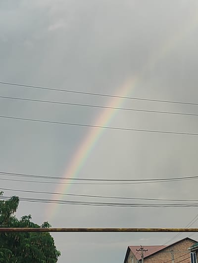 The image shows a dull, overcast sky in Tursunzoda, Tajikistan, with a faint rainbow arcing across the upper portion. Multiple dark power lines crisscross the frame horizontally. In the lower portion of the image, the top of a dense green tree is visible on the left, and the roofline and brick facade of a building are partially visible on the right. A thick, horizontal pipe or beam runs across the middle ground. No people or active interactions are depicted. The overall scene is one of a cloudy day with elements of urban infrastructure and nature.