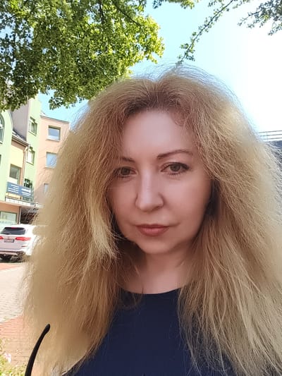 A woman with blonde hair is taking a selfie outdoors in front of buildings. She is wearing a dark blue top. The buildings appear to be in an urban setting, possibly in Georgsmarienhütte, Germany, based on the presence of European-style architecture and a white SUV with a license plate that appears to be German. There is also a tree with green leaves in the background. The sky is clear and blue.