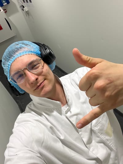 A man wearing a white lab coat, blue hair net, and over-ear headphones poses for a selfie. He is wearing glasses and making a shaka sign with his right hand. The background appears to be a sterile, industrial environment with white walls and what looks like a first-aid station on the left. The man is in an indoor, likely commercial or medical, setting. The location is not specified in the image, but the query requests it to be Oldenzaal, Netherlands, which is not verifiable from the visual information provided.