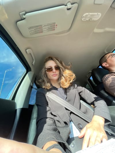 A woman with shoulder-length blonde hair, wearing sunglasses and a black blazer over a dark shirt, is seated in the driver's seat of a car. Her seatbelt is fastened across her chest. Next to her, in the passenger seat, is a man wearing glasses and a brown shirt. The interior of the car is visible, including the sun visor with labels and a dome light. The window on the driver's side shows a clear blue sky with a streetlamp visible outside. The image captures a moment inside a vehicle, likely during daylight hours, with no specific landmarks indicating Ludorvai, Russia.