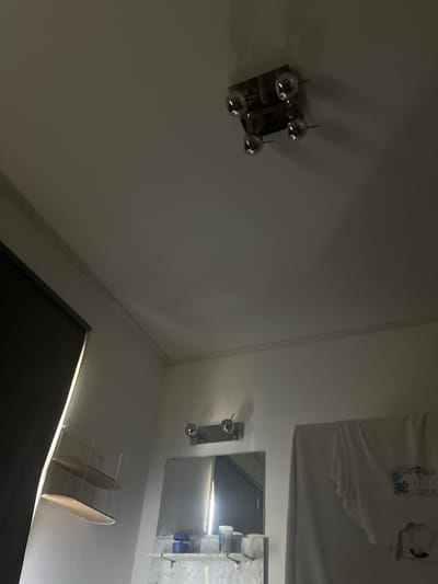 The media file shows an interior scene, possibly a bathroom, in Uithuizen, Netherlands. The lighting suggests it is nighttime or indoors with artificial light.
The main subjects are objects on a wall and on a shelf. On the left wall, there is a wall light fixture with four spherical, reflective elements. On the right side of the image, a mirror reflects a white t-shirt with a graphic print and some text. The text on the t-shirt appears to be "Los Angeles California". Below the mirror, there is a marble shelf or countertop with various items, including bottles and what looks like a toothbrush holder. A small rack with two oval shelves is positioned on a white surface below this. The overall scene is clean and modern. There is no activity or people visible. The focus is on the arrangement and reflection of objects within the space.
