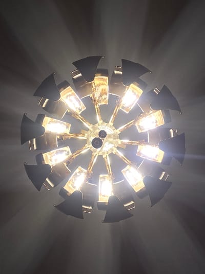 The image displays an illuminated circular ceiling light fixture, presented from a direct upward perspective. The fixture, reportedly located in Blainville-sur-Orne, France, consists of a central, lighter-toned hub from which multiple metallic, gold-toned arms radiate outwards. Each arm supports an incandescent or LED bulb, approximately 12 to 15 are fully or partially visible, all actively emitting a warm, bright light. Curved, dark grey or black, possibly metallic, shades partially enclose each bulb, directing the light. Distinct rays of light emanate from the illuminated bulbs, scattering into the surrounding dark grey environment, which is indicative of a ceiling or upper wall surface. No individuals, additional objects, or specific environmental identifiers beyond the fixture and its immediate background are present in the frame.