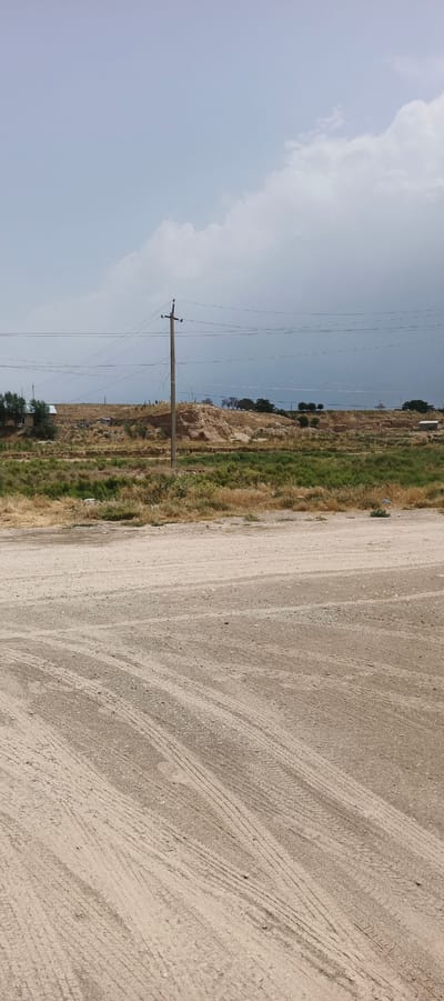 The image depicts an outdoor scene with a dirt road in the foreground. The road is dry and shows tire tracks. In the middle ground, there is a flat, overgrown area with dry grass and some green bushes. A prominent utility pole with multiple wires stretches across the scene, connected to other poles further in the distance. Behind this, there is a raised, eroded dirt mound with some structures visible on top. The background features more dry vegetation, scattered trees, and some small buildings. The sky is overcast with a mix of light and dark clouds, suggesting a potentially stormy or hazy day. The overall impression is a rural or semi-rural landscape.