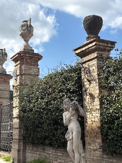 This daylight image captures a classical garden scene, likely part of an estate or public park, situated in Foligno, Italy. In the foreground, a weathered light-colored stone statue of a nude female figure stands, positioned slightly right of center. She holds a large bunch of grapes near her left shoulder with her raised left hand, while her right hand is positioned at her chest. A piece of drapery covers her lower right hip. The statue rests on a roughly textured base, possibly sculpted to resemble a tree stump.
Behind the statue, a dense, dark green trimmed hedge forms a continuous barrier across the mid-ground. Flanking the hedge on either side are two weathered stone gate pillars. The pillar on the left features a lower section of light-colored rough-hewn stone blocks, transitioning to reddish-brown bricks, topped by a decorative classical stone urn with Greek key patterns. To the immediate left of this pillar, a dark, ornate wrought-iron gate section is partially visible.
The pillar on the right mirrors the construction style of the left, with light stone blocks and reddish-brown bricks. It is crowned with a dark, textured, urn-shaped finial, possibly sculpted or overgrown. Both pillars have decorative wrought-iron brackets attached to their sides, with some sparse, dried-looking vines trailing around the right pillar.
The background consists of a clear blue sky interspersed with scattered white cumulus clouds. Strong sunlight illuminates the scene from the right, casting distinct shadows, particularly on the right pillar and hedge. A narrow strip of ground with patchy green vegetation is visible at the bottom of the image, immediately in front of a low brick wall that forms the base for the hedge and pillars.