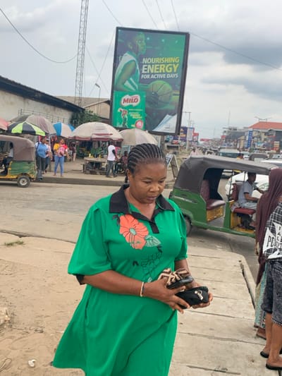 An adult Black woman with braided hair, wearing a green dress with a black collar and orange floral pattern, stands in the foreground on a dirt shoulder. She is holding a pair of dark sandals or slippers and possibly a small black pouch in her hands, her gaze directed downwards. A thin bracelet is visible on her right wrist.
The scene depicts a bustling urban street in Aba, Abia, Nigeria. To the woman's left, a paved road borders the shoulder, and further back, a green Keke Napep (motorized tricycle) is partially visible. Behind this, a vibrant open-air market unfolds, characterized by numerous individuals, market stalls with multi-colored umbrellas (white, green, blue), and a white building with a dark roofline in the background. Pedestrians, including a person in a blue shirt and another in a pink top, navigate this area.
To the woman's right, another green Keke Napep is positioned on the paved road, carrying a man in a light shirt. A second individual, partially obscured and wearing a brown headscarf and checkered top, stands near the Keke Napep. Beyond the road, a line of vehicular traffic extends down the street, flanked by various commercial buildings.
Prominently displayed above the market and street is a large billboard advertising "MILO," featuring a basketball player and the slogan "NOURISHING ENERGY FOR ACTIVE DAYS." Overhead, power lines crisscross against an overcast sky, and a tall metal communication tower is visible in the distance.