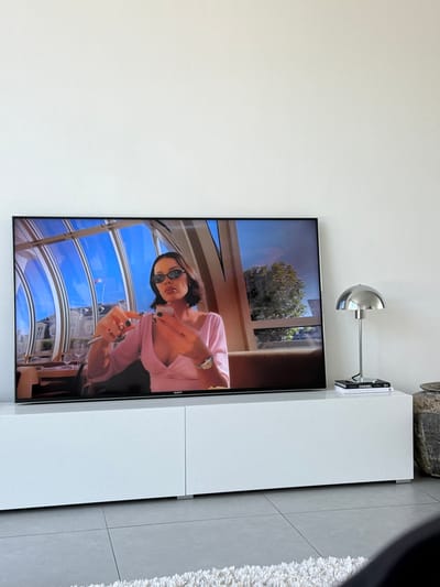 The image displays an indoor room located in Tielrode, Belgium. A large, black-framed Sony flat-screen television rests on a white, two-door media console. The television screen presents a still image of a female individual. This individual possesses dark hair, wears dark sunglasses, a light pink v-neck top, and dark nail polish. She holds a thin, dark object in her right hand, pointed upwards, while her left hand is open, fingers slightly spread, displaying a silver watch on her wrist. The scene depicted on the television is an upscale restaurant or cafe with large, arched glass windows revealing a clear blue sky, a white architectural building, and a French flag on an exterior pole. Green foliage is also visible outside the windows.
To the right of the television, a chrome-finished table lamp with a domed shade stands on the console. Below the lamp, two books are stacked; the uppermost book is labeled "CHANEL." A dark, textured pot or vase is partially visible on the far right. The room features white walls and light grey square floor tiles. A portion of a white shag rug is visible in the bottom foreground.
