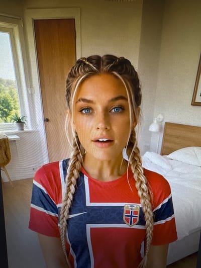 This media file is a close-up, vertically oriented photograph of a young woman wearing a red jersey with a blue and white design, featuring the Norwegian flag emblem on the chest. She has blonde hair styled in two braids that rest on her shoulders and chest. Her facial features are visible, including blue eyes and lips that are slightly parted, suggesting she is speaking or smiling. The background is softly blurred, but hints of an indoor setting are discernible, with what appears to be a bed and a wooden surface, possibly a door or furniture, in the upper part of the frame. A window with greenery visible outside is in the lower portion of the image. The lighting suggests it might be daytime, though no specific time is evident. There is no visible text in the image. The overall impression is that of a casual, personal snapshot.