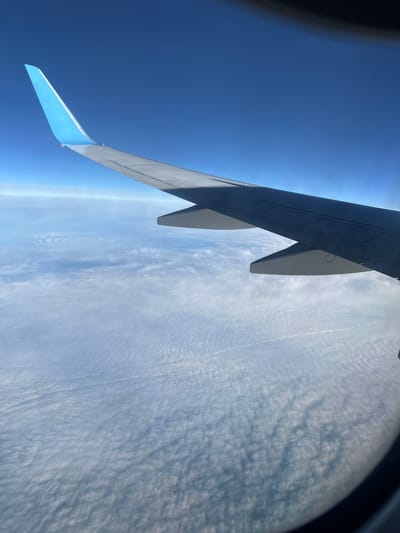 The image shows an airplane wing seen through an airplane window. The sky is visible above the wing and a layer of clouds is below the wing. There is no indication of Kitsjanovo, Russia, or any people, subjects, objects, or actions besides the wing, clouds, and sky seen from the airplane window.