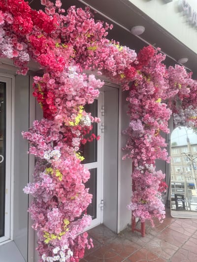 The image depicts a storefront in Achinsk, Russia, adorned with an abundant artificial floral arrangement. The branches, primarily composed of pink and red cherry blossoms with some yellow accents and small white daisy-like flowers interspersed, cascade over the entrance and down the side of the building. The storefront features a glass door with a white frame on the left and another glass door on the right, partially obscured by the floral display. A large, ornate mirror is positioned to the right of the entrance, reflecting a building with multiple windows and some Cyrillic signage. The ground is paved with reddish-brown tiles. The overall impression is one of a festive or decorative embellishment for the establishment.