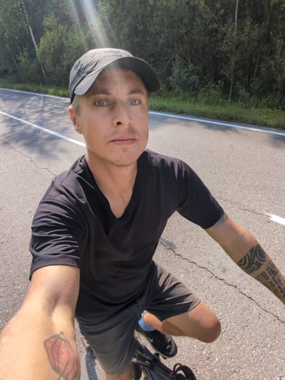 A man is riding a bicycle on a paved road. He is wearing a black t-shirt, black shorts, a black baseball cap, and black athletic shoes with blue socks. The man has blue eyes, fair skin, and light brown hair. He has tattoos on both arms. The road has a white line on the right side and appears to be cracked. On the left side of the road, there is a dense line of green trees and foliage, with some tall trees visible. The sky is bright and sunny, with some lens flare visible in the upper left corner of the image. The scene appears to be a rural or semi-rural road, likely in a forested area. The location is 17 Lineynyy Punkt, Russia.