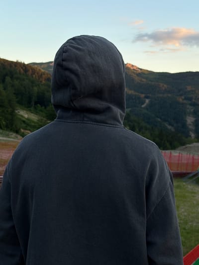 The image shows a person from behind, wearing a dark grey hooded sweatshirt, looking out at a mountainous landscape at dusk. The person is standing on a grassy area, with a red barrier visible in the background to the right. The mountains are covered in dark green trees, and the sky is a pale blue with hints of pink from the setting sun. The location is described as Katun Bačko brdo, Montenegro. The scene is peaceful and serene, with a sense of solitude.