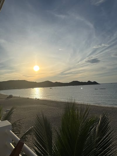 The image depicts a serene beach scene in Song Cau, Vietnam, during the late afternoon or early evening. The sun, a bright orb, is low on the horizon, casting a warm, golden glow across the sky and water. Wispy clouds streak across the blue sky, adding texture and depth.
In the foreground, lush green palm fronds are visible, framing the view of the beach and ocean. The sandy beach stretches along the coastline, appearing mostly empty except for a few scattered individuals and a parked vehicle on the left side. The ocean is calm, with gentle waves lapping the shore. Several small boats are visible scattered across the water, some anchored near the shore and others further out.
In the distance, rolling hills form a picturesque backdrop to the bay. The silhouette of these hills is softened by the descending sun. The overall atmosphere is peaceful and tranquil, evoking a sense of relaxation and natural beauty.