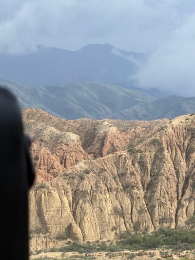 This image depicts a rugged, mountainous landscape with layers of hills and mountains receding into the distance. The foreground is dominated by reddish-brown and beige eroded hills, characterized by steep, ridged slopes and sparse vegetation. In the middle ground, greener, more rounded hills are visible, suggesting a transition in terrain. The background features distant mountains shrouded in a light blue haze and clouds, hinting at a vast, atmospheric expanse. A dark, blurry shape on the left side of the frame suggests the presence of an object or structure partially obscuring the view, possibly a vehicle or building from which the photograph was taken. The overall impression is one of natural, weathered topography under a diffused sky. This scene is consistent with the geological formations found in the vicinity of Tong, Kyrgyzstan, known for its canyons and eroded landscapes.