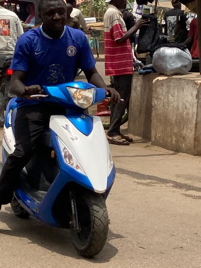 In Tudun Nupawa, Nigeria, a man wearing a blue Chelsea football club t-shirt sits on a blue and white scooter on a paved street. The man is looking at the camera. Several people stand behind the scooter, and objects like bags and clothes are visible. A concrete structure is to the side. The environment suggests a busy street scene.
