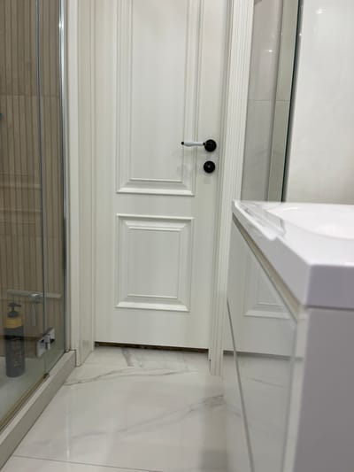 The image depicts a modern bathroom with white tiled flooring and a white paneled door. To the left of the door is a glass shower enclosure with ribbed tiled walls behind it, and a dark colored soap dispenser attached to the enclosure. The door has black hardware, including a knob and a lock. To the right of the door, a white countertop is visible, likely part of a vanity. The overall impression is of a clean and contemporary bathroom space. The location of Нясвіжскі сельскі Савет, Belarus is a rural area, so this scene likely depicts a residential bathroom.
