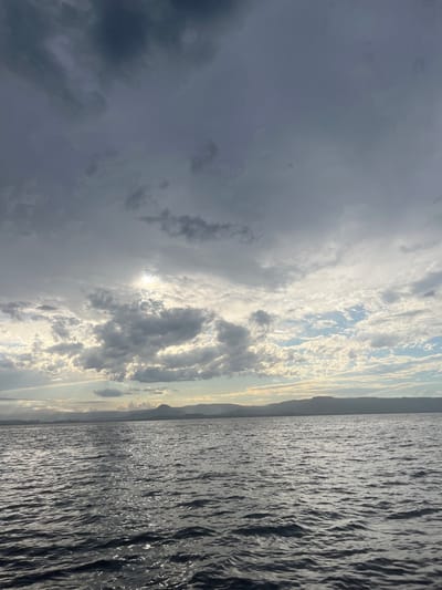 The image shows a landscape divided between the sky and the sea. The sky is dominated by dark and light gray clouds, with the sun trying to break through in the center. A distant, hazy shoreline is visible between the clouds and the water. The sea is dark and reflects the light, with visible ripples and waves across its surface. The image is oriented vertically.