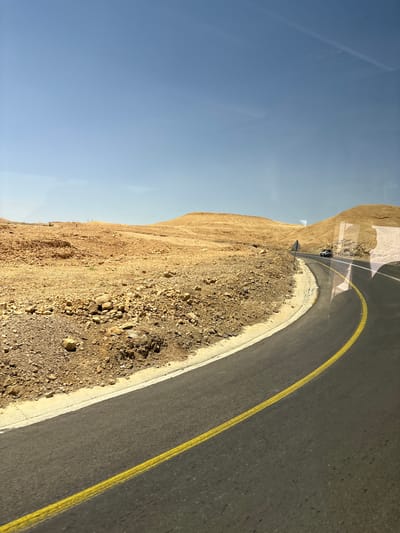 The image depicts a desert landscape with a winding asphalt road in the foreground. The road has a yellow line marking its center. To the left of the road, the terrain is rocky and sandy, with low, barren hills in the background. The sky is clear and blue. A white SUV is visible driving on the road in the distance. The scene appears to be a scenic route through the Arava Tichona Regional Council, Israel, characterized by its arid and mountainous terrain.
