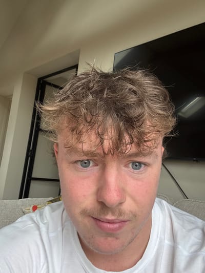 A male subject is depicted in an indoor residential or commercial setting located in Skipsea, United Kingdom. The subject, a young adult male with fair skin, light blue eyes, and light brown, messy, curly hair, is positioned centrally, looking directly at the camera. He has a light brown mustache and visible stubble. His skin exhibits some redness around the nose and cheeks. He is wearing a plain white t-shirt. The background on the left reveals a section of a dark-framed internal structure, possibly a doorframe or shelving unit, against a light-colored wall. On the right, a large, dark, rectangular object, likely an inactive television screen, is visible. A light-colored, textured fabric surface, consistent with a sofa or cushion, is partially visible in the lower-left portion of the frame. The upper background features a light, untextured wall or ceiling.
