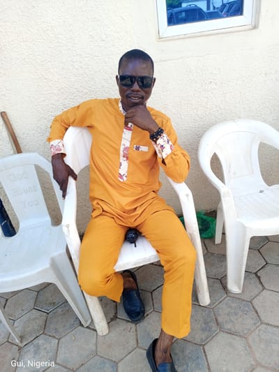 An adult male wearing sunglasses and a bright orange traditional outfit sits on a white plastic chair. He is leaning back and resting his chin on his hand, with his left arm propped on the back of the chair. The outfit has white accents with red and pink floral patterns on the collar and cuffs. He is also wearing dark-colored beaded bracelets on his left wrist and black leather loafers. A black walkie-talkie is visible on the seat of the chair between his legs. The background features a textured cream-colored wall and a window with reflections of vehicles. To his left and right are two more white plastic chairs, one with the inscription "God Bless You." The ground is paved with hexagonal tiles. The image is labeled "Gui, Nigeria."