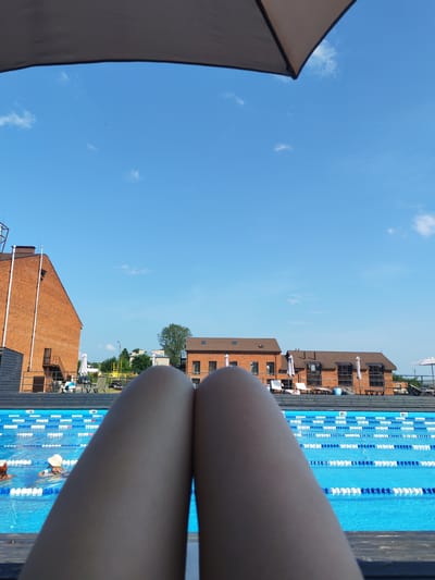 The image depicts a person's legs in the foreground, positioned in front of a swimming pool with lane markers. The pool area is bordered by wooden decking and lounge chairs. In the background, there are brick buildings with windows and tiled roofs, suggesting a resort or residential complex. Trees and a clear blue sky with a few clouds complete the outdoor scene. There are two individuals visible in the swimming pool, one wearing a white hat and another with their head submerged. The perspective suggests the photo was taken from a reclining position, possibly on a lounge chair, under a large umbrella that partially obscures the top of the frame. The location is identified as Pirogovo, Russia.