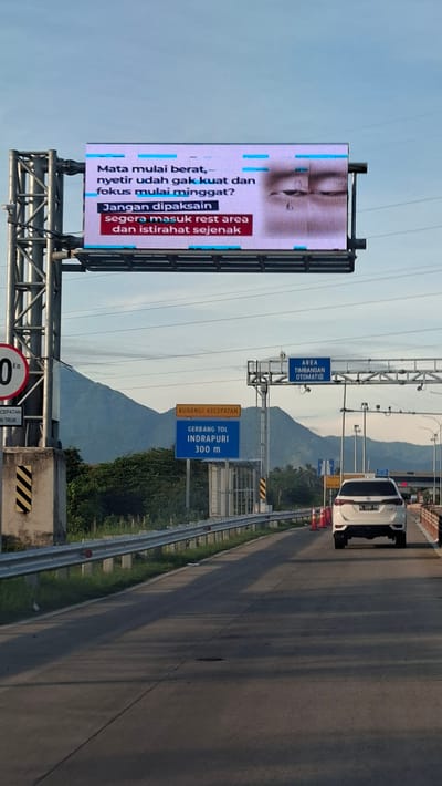 The image shows a digital billboard on a highway with a message about driver fatigue. The billboard displays text in Indonesian that translates to "Eyes starting to feel heavy, already can't stand driving and focus is starting to waver? Don't force it, immediately enter a rest area and rest for a while." The background of the billboard features a close-up image of a person's tired eyes.
Below the billboard, there are road signs indicating "KURANGI KECEPATAN" (Reduce Speed) and "GERBANG TOL INDRAPURI 300 m" (Indrapuri Toll Gate 300 m). There is also a sign for "AREA TIMBANGAN OTOMATIS" (Automatic Weighing Area).
The highway itself is visible, with a white SUV driving on the right lane. The environment appears to be mountainous with some vegetation. The sky is clear and bright, suggesting it's daytime. The location is identified as Sihomlhok, Indonesia.