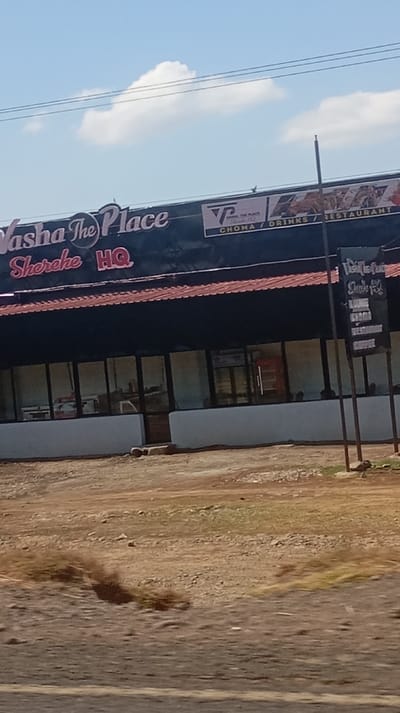 A roadside restaurant called "Vasha The Place Sherehe HQ" features a long, low-profile building with a dark roof and many windows. A sign advertises "Choma/Drinks/Restaurant" alongside a menu board listing "Lounge," "Choma," "Restaurant," and "Coffee." The exterior has a simple, somewhat unfinished look with a dusty ground area in front. A few birds are perched on the roof of the building. Overhead, electrical wires are visible against a bright blue sky.