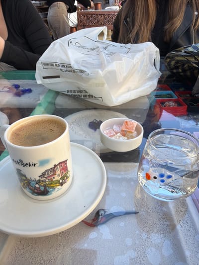 The image captures a vibrant tabletop scene, likely outdoors or in a well-lit cafe, with a person partially visible in the frame. A white ceramic cup of foamy Turkish coffee rests on a matching saucer, featuring a colorful illustration of buildings and the word "Eskişehir" printed on its side, confirming the location as Eskişehir, Turkey. Adjacent to the coffee, a small white bowl holds several cubes of colorful Turkish delight (lokum) in orange and light pink hues.
To the upper right, a clear glass ornament resembling a fish, with distinctive blue and orange accents, is visible. A slender, decorative bird figurine, possibly made of ceramic or wood, also sits on the table. A white plastic shopping bag, partially crumpled, occupies a significant portion of the left side of the table. The bag displays text in Turkish, including "OĞUZARİ PAŞA MH. PAZARNALO TEL: 0546 205 05 60" and "PAŞA MH. TEL: 0546 20," which likely denote a business name, address, and phone number.
The background shows glimpses of the person's long brown hair, part of a black jacket, and a lower leg in black trousers, suggesting a casual setting. The table surface appears reflective, capturing ambient light and faint reflections of other objects. Bright, natural lighting indicates it is daytime. The overall scene suggests a leisurely moment, enjoying traditional Turkish refreshments.