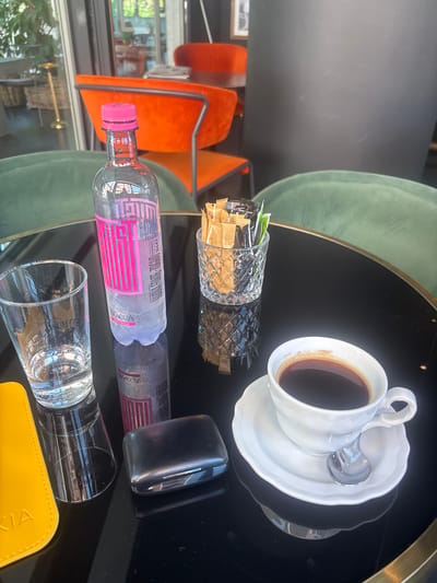The image displays a table at what appears to be an outdoor cafe in Bergamo, Italy. The scene is captured from a top-down perspective, with the objects on the table arranged in a linear fashion.

The prominent objects include a white ceramic cup of black coffee, resting on a saucer with a small silver spoon. Beside the coffee cup are two glass containers filled with sugar packets. Further along the table are two bottles of water, one partially obscured by the other. The bottle in the foreground has a pink cap and a label with pink lettering that reads "ACQUA FONTE - ZUCCONI". The other water bottle has a similar label with "BRACCA" visible. To the right of the water bottles is a small, dark rectangular object, possibly a phone or a small case. Finally, a clear glass filled with water or a clear beverage sits in the foreground, partially covering a yellow leather item.

The background reveals glimpses of the cafe's setting, with plush, green velvet chairs visible on the left and vibrant orange chairs on the right. Through a large window, a street scene with trees and buildings can be seen, suggesting daytime and fair weather. The overall ambiance appears relaxed and casual, indicative of a typical cafe experience. No people are visible in the frame.