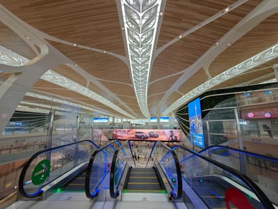 The image depicts the interior of what appears to be an airport or transportation hub in the Baiyun District of China. The perspective is from the top of an escalator, looking down towards the main floor. Two escalators with blue lights on the sides are visible. Glass barriers and railings surround the escalators.
In the background, there is a large advertising display promoting "ID.ERA 9X" Volkswagen vehicles. Beyond the advertising display are people moving about in the main area.
The architectural design of the ceiling is a prominent feature, with wooden slats and white supports, creating an intricate pattern with a central line of decorative lighting resembling leaves. There are also multiple flat screen displays and artificial green walls in the vicinity.
Signage in Chinese characters and international symbols is also present.