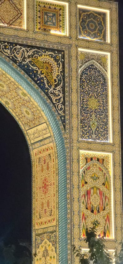 The image shows a detailed view of an ornately decorated archway, likely a monument or an entrance, in Satkak, Uzbekistan. The archway is adorned with intricate geometric patterns and floral motifs rendered in vibrant colors such as blue, yellow, green, red, and white. The decorative elements appear to be ceramic tiles or mosaics, possibly illuminated from within in some sections, creating a glowing effect.
On the left side of the arch, there is text inscribed within a yellow rectangular panel. The text reads: "The light of knowledge, the beacon of hearts." This inscription is in English.
Below this inscription and further along the curve of the arch, there is a panel featuring a repeating pattern of stylized motifs, possibly representing paisley shapes or teardrops, in red, orange, yellow, and green against a lighter background.
The right side of the archway features several rectangular panels, each with distinct, elaborate patterns. One prominent panel contains a stylized, star-like floral design in shades of green and blue on a dark blue background. Another panel, situated below it, showcases a more complex design with vertical bands of vibrant colors, including red, pink, yellow, and green, interspersed with white swirling patterns. The overall impression is one of rich artistry and cultural heritage, characteristic of Central Asian architecture. The background is dark, suggesting the image was taken at night or in a dimly lit environment.