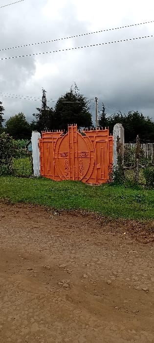 The image depicts a closed, ornate orange metal gate marking an entrance from a dirt road in Gatakaine, Kenya. The gate, centrally positioned, features two main panels with a distinctive oval shape and intricate scrollwork, flanked by vertical bars. It is supported by two rough-textured, light-colored posts; the left post is a solid white structure, while the right post appears to be a narrower, possibly concrete or stone pillar, partly obscured by vegetation and a weathered wooden fence structure. A strip of green grass and low-lying plants separates the gate from the uneven, reddish-brown dirt road in the foreground, which shows signs of vehicle tracks and small stones. In the background, beyond the gate and vegetation, a dense line of dark green trees stands against an overcast, grey sky. Three parallel utility lines are visible stretching across the upper portion of the sky. No individuals are present in the scene.