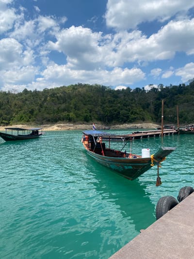 The image depicts a longtail boat docked at a pier in Ban Ta Khun, Thailand. The boat is dark green and has a white container on board, with yellow tassels hanging on the bow. The boat is tied to a grey pier with a rope. Old tires are used as bumpers on the side of the pier. The water is a turquoise color. In the background is a wooded shoreline. The sky is blue with white clouds. Another boat is partially visible in the lower portion of the image.